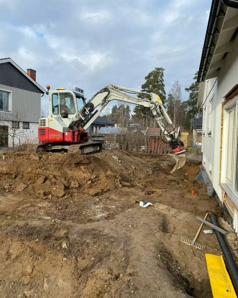 Grävmaskin vid ett öppet schakt intill ett hus