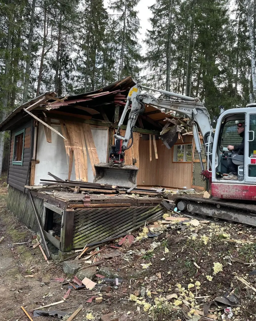 Grävmaskin river en äldre stuga i skogsmiljö